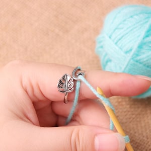 Può includere: Un anello in metallo color argento con un design a foglia di monstera su un'estremità e un gancio curvo sull'altra, indossato su un dito mentre si guida un filo di lana azzurro con un uncinetto in legno per facilitare il lavoro a maglia.