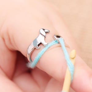 May include: A silver-colored ring featuring a dachshund dog design. The ring is wrapped around a finger with light blue yarn and a wooden crochet hook. The dog has a light blue accent on its back.