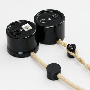 May include: Two cylindrical black wall sockets with glossy finishes. One unit features a white and a black modular port, while the other has two circular coaxial ports. Both are connected to a twisted beige fabric cord with black plastic components.