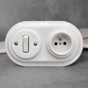 May include: A white ceramic wall plate featuring an oval shape with a single toggle light switch on the left and a circular electrical socket on the right. The switch and socket are integrated into a smooth, glossy, continuous white housing.