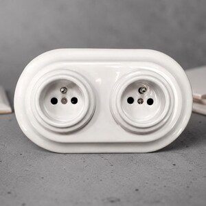 May include: A white ceramic double electrical wall socket featuring a smooth, rounded, oval-shaped faceplate. The unit includes two recessed circular sockets, each with two pin holes and a central grounding pin, designed for standard electrical connections.