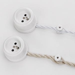 May include: Two white ceramic electrical outlets with three-prong sockets, each connected to a twisted fabric cord. One cord is white, the other is beige. The cords are secured with small ceramic fasteners.