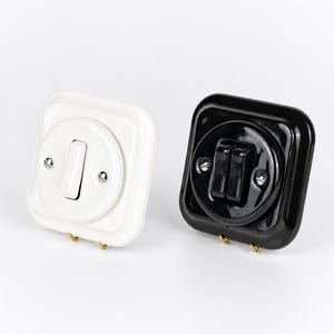 May include: Two vintage-style light switches. One is white with a toggle switch, the other is black with a double-button design. Both have a square base and are mounted with brass screws, ideal for home decor.