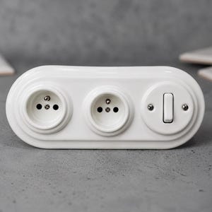 以下が含まれることがあります： 2つの円形コンセントと1つの長方形の照明スイッチを備えた白いセラミック製の壁プレート。細長い楕円形の器具は光沢のある仕上げで、スイッチ機構を固定する金属製のネジが見えています。