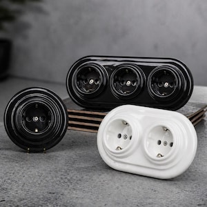 May include: A selection of ceramic electrical sockets in black and white. The sockets have a retro design. One is round, while others are oval and hold two or three sockets. The sockets are displayed on a grey surface. Measurements are in centimetres.