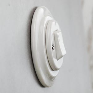 May include: A white ceramic light switch with a circular base plate and a rectangular toggle lever. The switch features a glossy finish and a single visible metal screw securing the central mechanism to the round mounting plate.