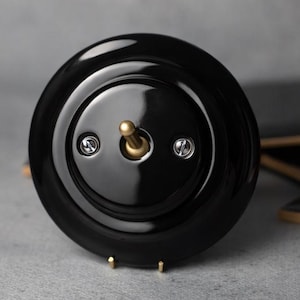 May include: A glossy black light switch with a circular design. The switch features a brass toggle and two small screws. The switch is mounted on a gray surface, with a few black tiles in the background.