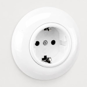 Può includere: Una presa elettrica a muro circolare di colore bianco con una finitura liscia e lucida. La presa presenta due fori rotondi, una vite centrale e due clip metalliche di messa a terra posizionate nella parte superiore e inferiore.