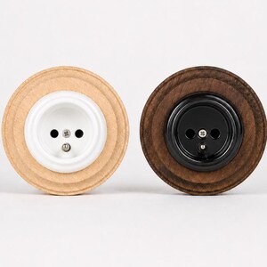 May include: Two circular electrical wall outlets with wooden mounting plates. One outlet features a light-colored wood frame with a white center, while the other has a dark-colored wood frame with a black center. Both have two pin holes.