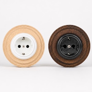 May include: Two circular electrical wall outlets with wooden frames. The left outlet features a light-toned wood frame with a white center, while the right outlet has a dark-toned wood frame with a black center. Both have two pin holes.