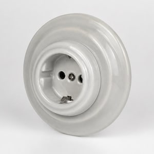 Può includere: Una presa elettrica a muro rotonda in ceramica grigio chiaro con finitura lucida. L'unità presenta una piastra di base circolare con un design a gradini e un'area centrale incassata contenente due fori per i pin e una vite centrale.