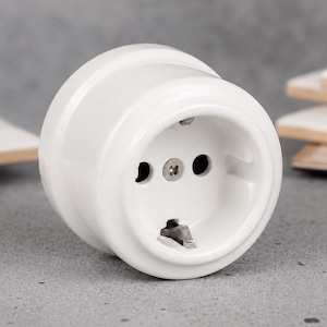 May include: A white ceramic electrical wall socket with a round profile. The socket features two circular pin holes, a central metal screw, and a metal grounding clip at the bottom, designed for standard electrical plug connections.
