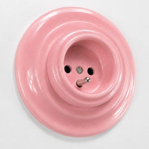 May include: A circular, light pink electrical wall socket with a glossy finish. The socket features a stepped, concentric design with a central grounding pin and two circular openings for electrical plugs, all in a matching light pink colour.