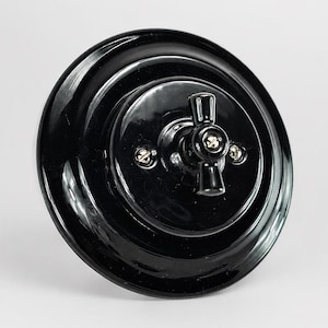 May include: A round, glossy black light switch with a stepped, circular base and a central rotating knob. The switch features a small, cross-shaped handle and is secured with two visible metal screws on the face, presenting a smooth, reflective finish.