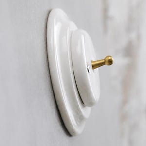 May include: A white ceramic light switch with a tiered, circular base and a small, protruding brass-colored toggle lever. The switch features a smooth, glossy finish and is mounted flush against a flat, neutral-colored wall surface.