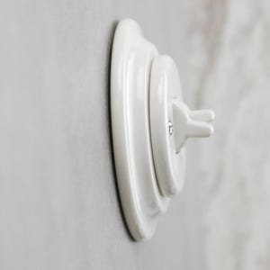 May include: A glossy white ceramic light switch with a round base plate and a protruding toggle lever. The switch features a stepped, circular design with a smooth, reflective finish and a small metal screw visible on the central toggle mechanism.