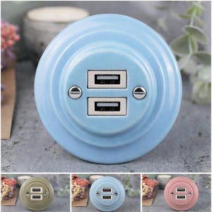 Puede incluir: Un cargador USB de cerámica azul claro con dos puertos USB. El cargador tiene un diseño circular con un anillo exterior ligeramente elevado y está asegurado con dos tornillos plateados.