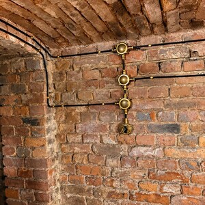 May include: A brick wall and ceiling with black electrical wiring and brass fixtures. The wiring runs along the ceiling and down the wall, connecting to the brass fixtures. The brick is a mix of red, brown, and grey.