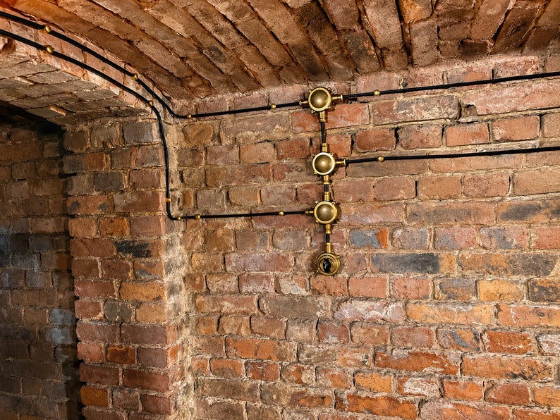 May include: A brick wall and ceiling with black electrical wiring and brass fixtures. The wiring runs along the ceiling and down the wall, connecting to the brass fixtures. The brick is a mix of red, brown, and grey.