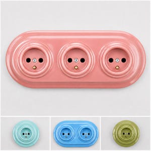 May include: A triple electrical wall socket plate in a glossy pink finish with three circular outlets, each featuring two pin holes and a central screw. The design is oval-shaped with concentric rings surrounding each individual socket opening.