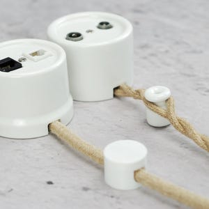 May include: Two white cylindrical wall-mounted electrical junction boxes with ports for data cables. One box features a black and a white modular jack, while the other has two circular ports. Both are connected to twisted beige fabric-covered cords.