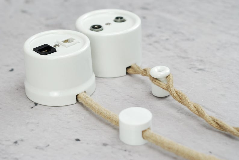 May include: Two white cylindrical wall-mounted electrical junction boxes with ports for data cables. One box features a black and a white modular jack, while the other has two circular ports. Both are connected to twisted beige fabric-covered cords.