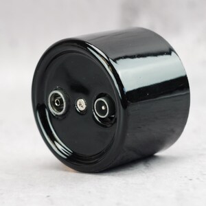 May include: A cylindrical black ceramic component with a glossy finish. The circular face features two circular electrical ports positioned on opposite sides of a central silver-colored screw, providing a functional interface for electrical connections.