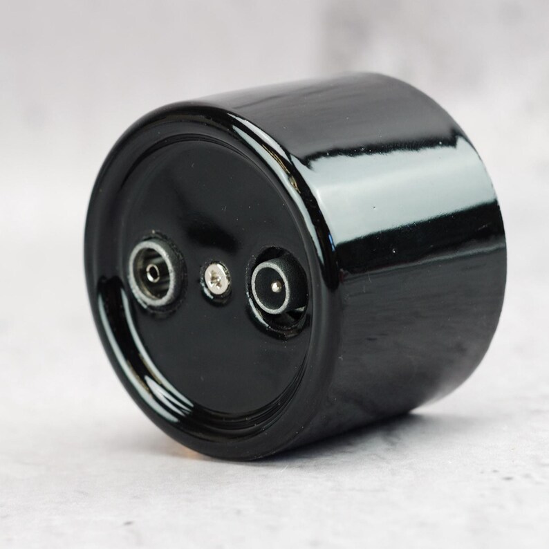 May include: A cylindrical black ceramic component with a glossy finish. The circular face features two circular electrical ports positioned on opposite sides of a central silver-colored screw, providing a functional interface for electrical connections.