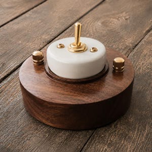 Può includere: Una base circolare in legno marrone scuro con un componente centrale in ceramica bianca e un interruttore a levetta in ottone. Due piccole viti in ottone fissano il pezzo in ceramica e due manopole in ottone sono sulla base in legno.
