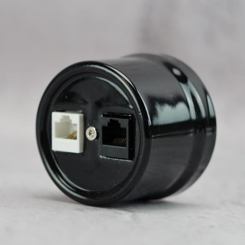 May include: A cylindrical black wall plate featuring a glossy finish with one white RJ11 telephone jack and one black RJ11 telephone jack positioned side by side, secured by a central silver screw on the circular face of the device.