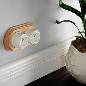 May include: A double electrical wall outlet featuring two white cylindrical sockets mounted on a light-toned wooden oval plate. The unit is installed horizontally on a smooth gray wall, positioned above a white baseboard molding.