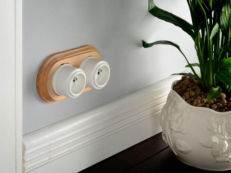 May include: A double electrical wall outlet featuring two white cylindrical sockets mounted on a light-toned wooden oval plate. The unit is installed horizontally on a smooth gray wall, positioned above a white baseboard molding.