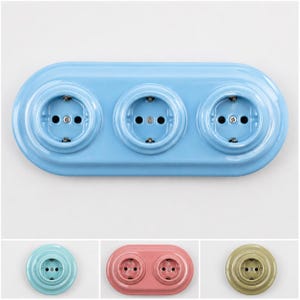 May include: A light blue triple electrical wall outlet with a glossy finish and a rounded rectangular base. Below, three additional outlets are shown: a single light blue outlet, a double pink outlet, and a single olive green outlet.