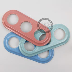 May include: Three elongated metal switch plates with rounded edges, each featuring three circular cutouts. The plates are finished in glossy pastel pink, light blue, and a darker blue. A circular logo with the text SOCKET AND SWITCH is visible.