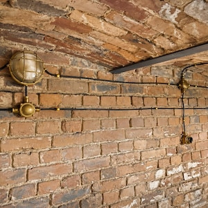 May include: A brick wall with exposed electrical wiring and brass light fittings. The wall is made of red and brown bricks, and the ceiling is arched. The light fittings have a cage-like design, giving a vintage feel.