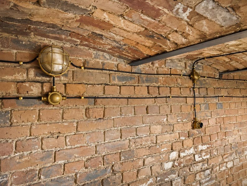 May include: A brick wall with exposed electrical wiring and brass light fittings. The wall is made of red and brown bricks, and the ceiling is arched. The light fittings have a cage-like design, giving a vintage feel.