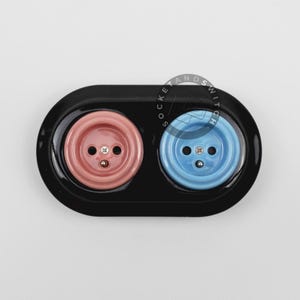 May include: A black oval double electrical wall outlet plate featuring one pink and one light blue circular socket insert. The center of each socket has two pin holes and a small metal screw. The text SOCKET AND SWITCH is printed in a circle.