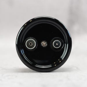 May include: A circular black electronic component featuring a glossy finish. The face displays two recessed circular ports positioned symmetrically on either side of a central silver-colored Phillips head screw, all set within a smooth black frame.