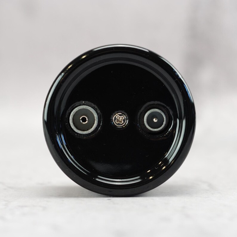 May include: A circular black electronic component featuring a glossy finish. The face displays two recessed circular ports positioned symmetrically on either side of a central silver-colored Phillips head screw, all set within a smooth black frame.