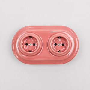 May include: A glossy, oval-shaped double electrical wall outlet cover in a solid dusty pink color. The unit features two circular recessed sockets with metallic screws in the center, designed for standard electrical plug connections.
