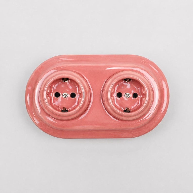 May include: A glossy, oval-shaped double electrical wall outlet cover in a solid dusty pink color. The unit features two circular recessed sockets with metallic screws in the center, designed for standard electrical plug connections.