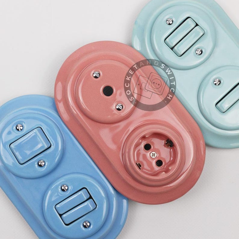 May include: Three oval-shaped wall plates with electrical components in light blue, dusty pink, and pale turquoise. Each plate features a toggle switch and a circular outlet, secured with small metal screws. A circular logo reads SOCKET and SWITCH.