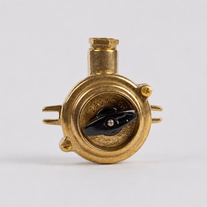 May include: A brass-colored valve with a black handle and a small, round dial. The valve has a circular body with two small, horizontal extensions and a threaded top. The background is a plain, light gray.