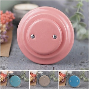 Może przedstawiać: Różowy ceramiczny świecznik o okrągłym kształcie i dwóch srebrnych śrubach. Świecznik ma warstwowy wygląd i jest pokazany z trzema innymi wariantami kolorystycznymi: niebieskim, beżowym i innym odcieniem niebieskiego.