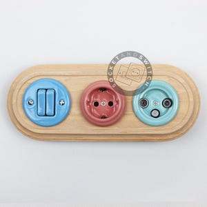 May include: An oval light-colored wooden wall plate featuring three circular components: a light blue double toggle light switch, a dusty pink electrical power outlet, and a light turquoise triple-port television and data connection socket.