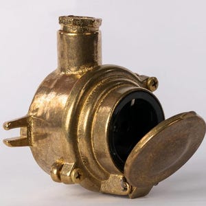 May include: A vintage brass electrical junction box with a hinged cover. The box has a circular body with a black interior and is secured with screws. The brass has a weathered appearance.