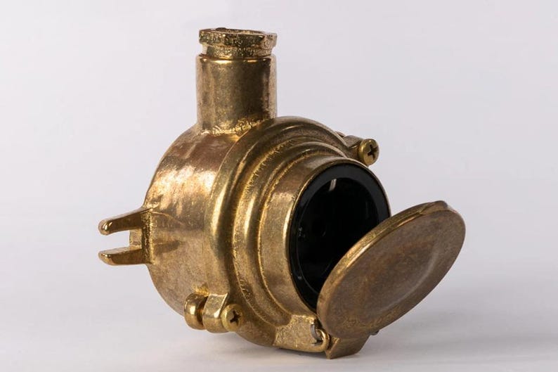 May include: A vintage brass electrical junction box with a hinged cover. The box has a circular body with a black interior and is secured with screws. The brass has a weathered appearance.