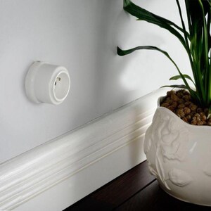 May include: A white circular electrical wall outlet mounted on a smooth white wall surface. The outlet features a recessed center with two small circular contact points and a rounded exterior casing, positioned above a white baseboard trim.