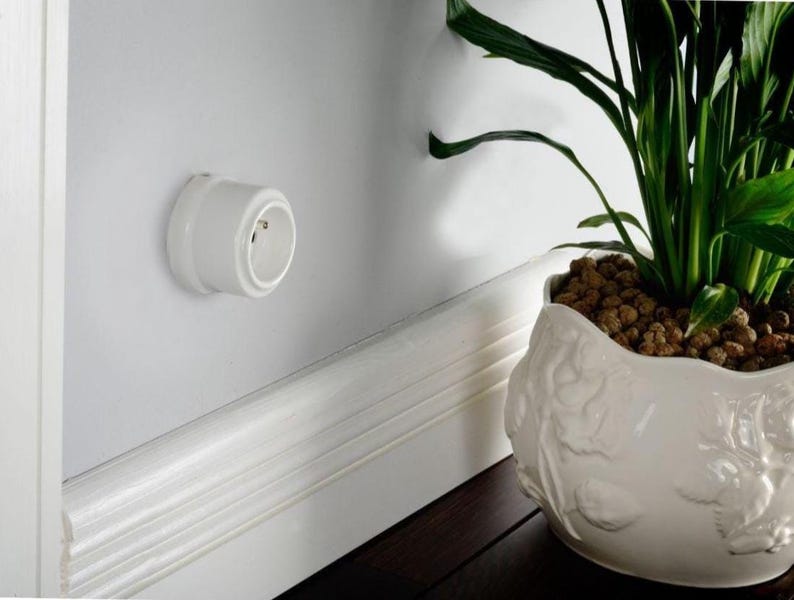 May include: A white circular electrical wall outlet mounted on a smooth white wall surface. The outlet features a recessed center with two small circular contact points and a rounded exterior casing, positioned above a white baseboard trim.