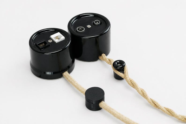 May include: Two cylindrical black wall sockets with glossy finishes. One unit features a white and a black modular port, while the other has two circular coaxial ports. Both are connected to a twisted beige fabric cord with black plastic components.